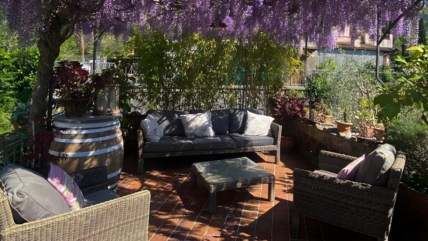 Een terras met een bank, rieten stoelen en een grote plantenbak.