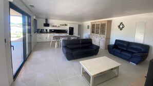 Et moderne opholdsområde med sofa, sofabord og spiseområde med stole samt et køkken i baggrunden.