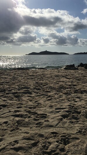 Ein sandiger Strand mit Fußspuren, die zum Wasser führen, eine entfernte Insel und ein teilweise bewölkter Himmel.