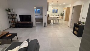 Ein modernes Wohnzimmer mit Sofa, Fernseher und Unterhaltungseinheit. Ein Essbereich mit Küche im Hintergrund.