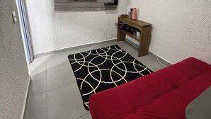 Una habitación con un televisor montado en la pared, un sofá rojo, un tapete blanco y negro estampado y un estante de madera con utensilios de cocina.