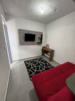 A room with a television mounted on a wall, a red sofa, a black and white patterned rug, and a wooden shelf with kitchen items. - Furnished 1-Bedroom Apartment in the North Zone (São Paulo)