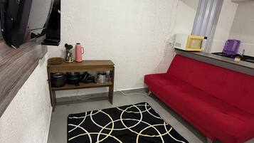 A compact living space with a red sofa, a black and gold patterned rug, a wooden shelf with kitchen items, a microwave, and a television.