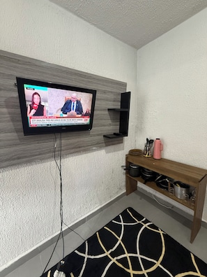 En tv som är monterad på en vägg med en intressant yta, en trähylla med köksredskap och en svartvit, mönstrad matta.