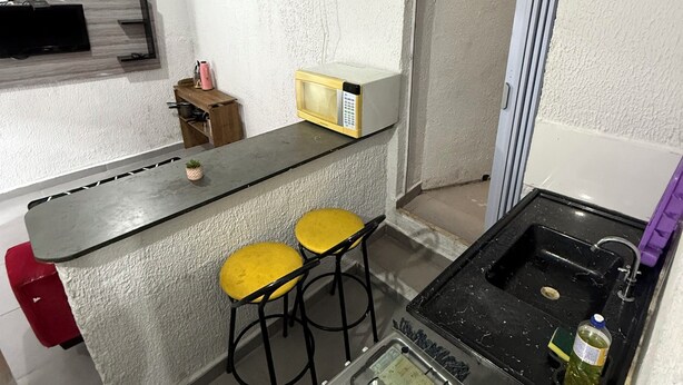 Cocina compacta con estufa a gas, microondas, dos taburetes amarillos, un lavamanos pequeño y un televisor montado en la pared.