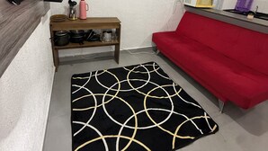 Un espacio habitable compacto con un sofá rojo, un tapete negro y dorado, una estantería de madera con utensilios de cocina, un microondas y un televisor sobre un mueble.