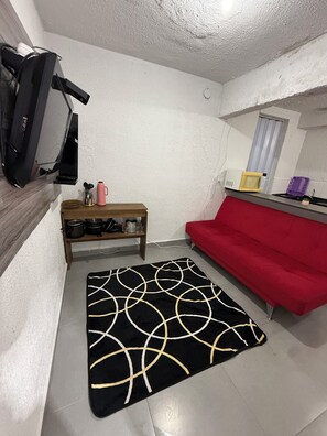 A compact living space with a red sofa, a black and gold rug, a wooden shelf with kitchen items, a microwave, and a TV on a stand. - Furnished 1-Bedroom Apartment in the North Zone (São Paulo)
