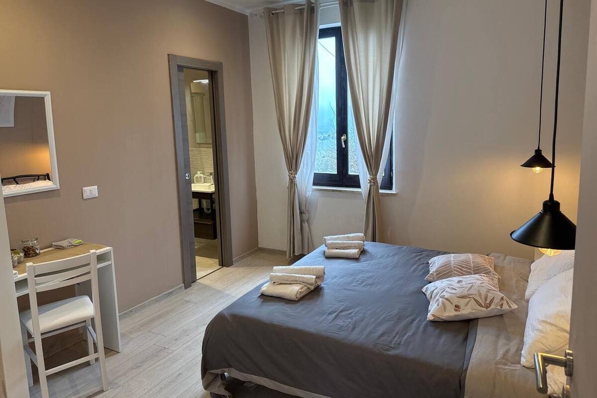 A bedroom with a bed, a desk, a chair, a window with curtains, and a hanging light fixture.