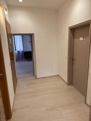 Un pasillo con suelo de madera, una puerta a la derecha y otra puerta entreabierta a la izquierda que da a otra habitación.