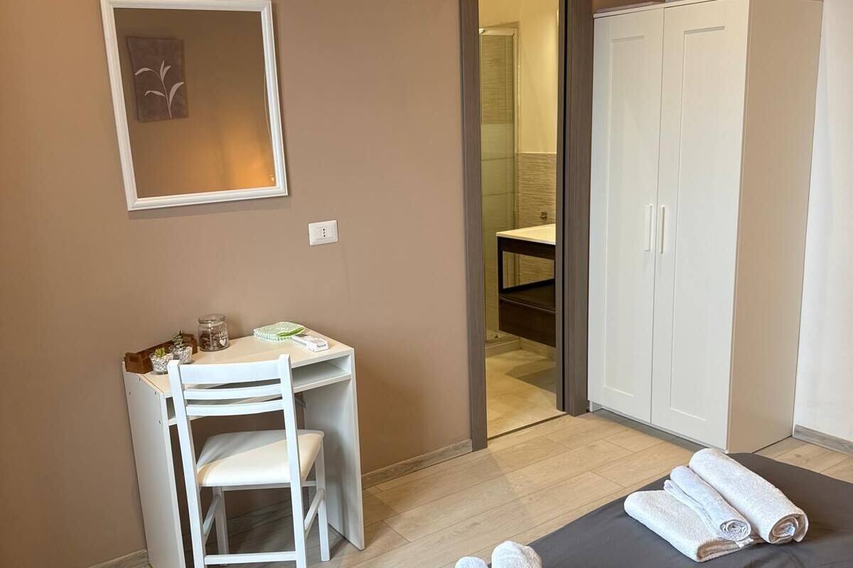 A hotel room with a bed, a desk with a chair, a wardrobe, and a bathroom visible through an open door.