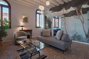 En moderne stue med sofa, lænestol, glasbord og et træmotiv på væggen.