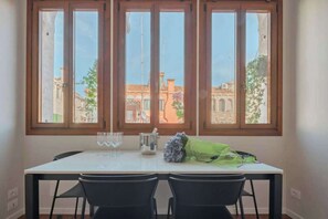 Ein Essbereich mit Tisch und Stühlen, große Fenster mit Blick auf Gebäude und ein Blumenstrauß auf dem Tisch.