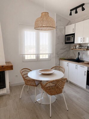 Eine moderne Küche mit weißen Schränken, einem runden Esstisch mit Korbstühlen, einer hängenden Rattan-Leuchte und einem großen Fenster.
