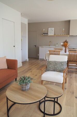 Un salon moderne avec un canapé, deux fauteuils, une petite table sur laquelle est posée une plante, et une cuisine en arrière-plan.