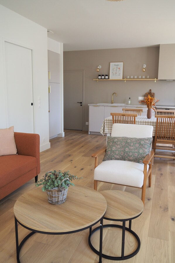 Ein modernes Wohnzimmer mit einer Couch, zwei Sesseln, einem kleinen Tisch mit Pflanze und einer Küche im Hintergrund.