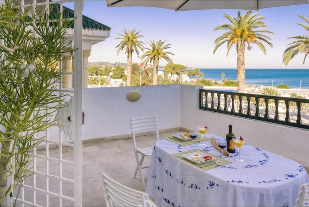 Een balkon met een eettafel voor twee, met uitzicht op een strand met palmbomen en de zee.