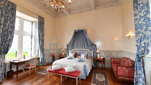 Un dormitorio con cama con dosel, un banco rojo, un sofá rojo, una araña y vistas al exterior con vegetación.