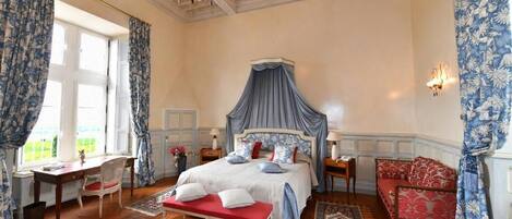 A bedroom with a four-poster bed, a red bench, a red sofa, a chandelier, and a view of greenery outside.