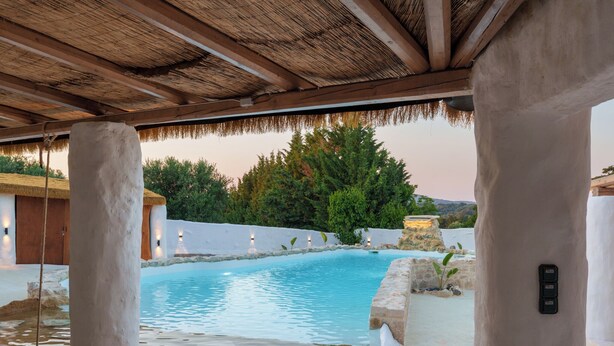 A pool area with a thatched roof, stone pillars, and a view of greenery and hills. - Aspasia Wabi Retreat by CVR (Roustika)