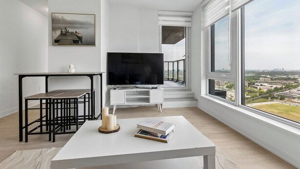 A modern living room with a large window, a flat-screen TV, a dining table, and a bookshelf. - Stylish 2bed2bath| Parking | Gym | Sauna|4ppl (Toronto)
