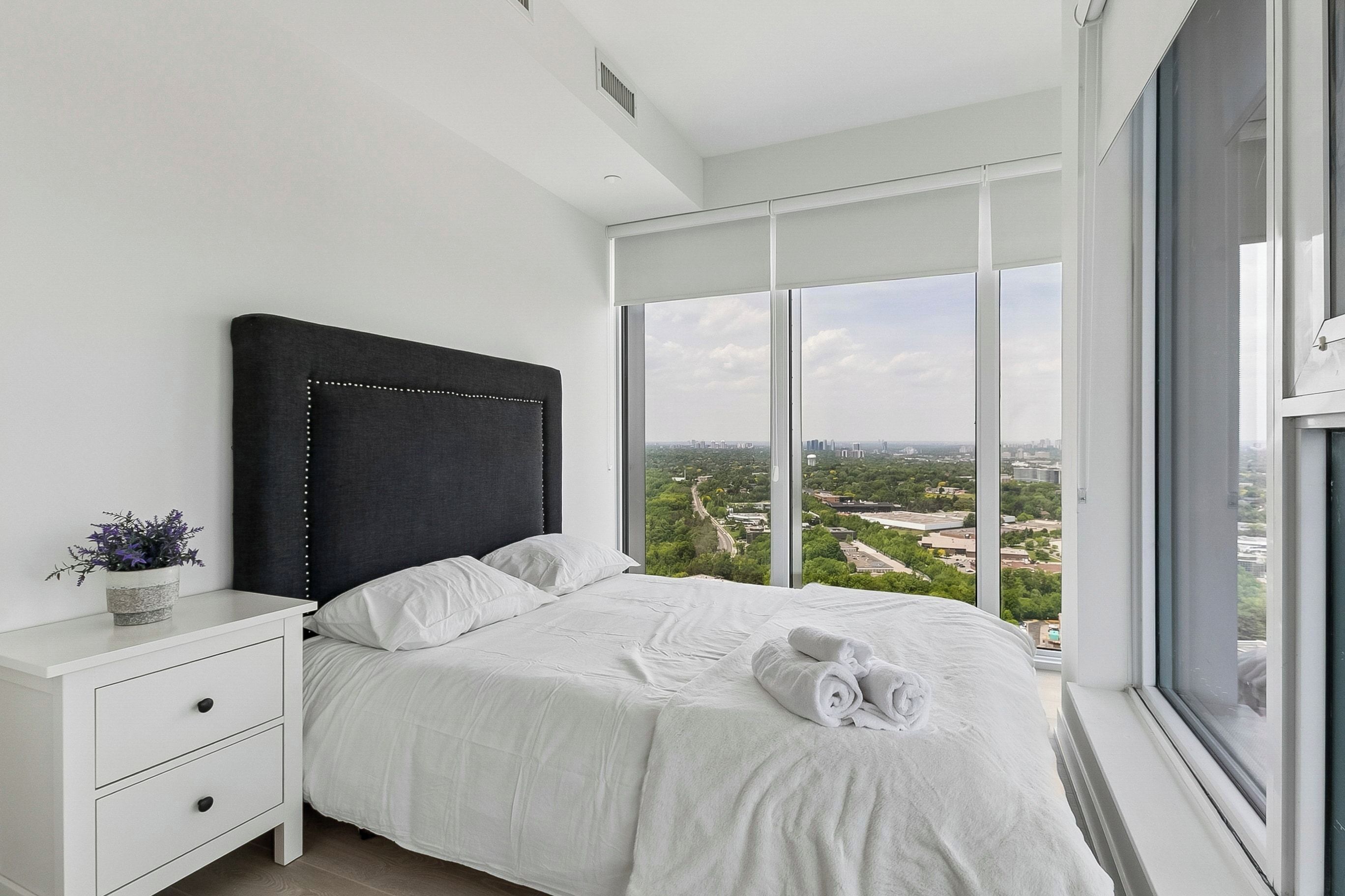 Quarto moderno com uma cama grande, uma mesa de cabeceira e vista para a cidade através de janelas do chão ao teto.