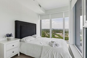 A modern bedroom with a large bed, a nightstand, and a view of the city through floor-to-ceiling windows.
