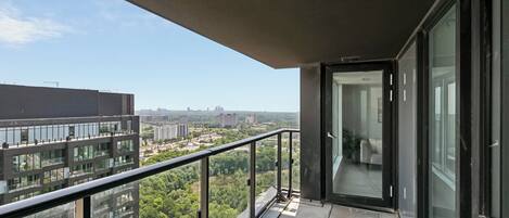 A balcony with a glass door leading to an interior space, a metal railing, and a view of urban buildings and greenery.