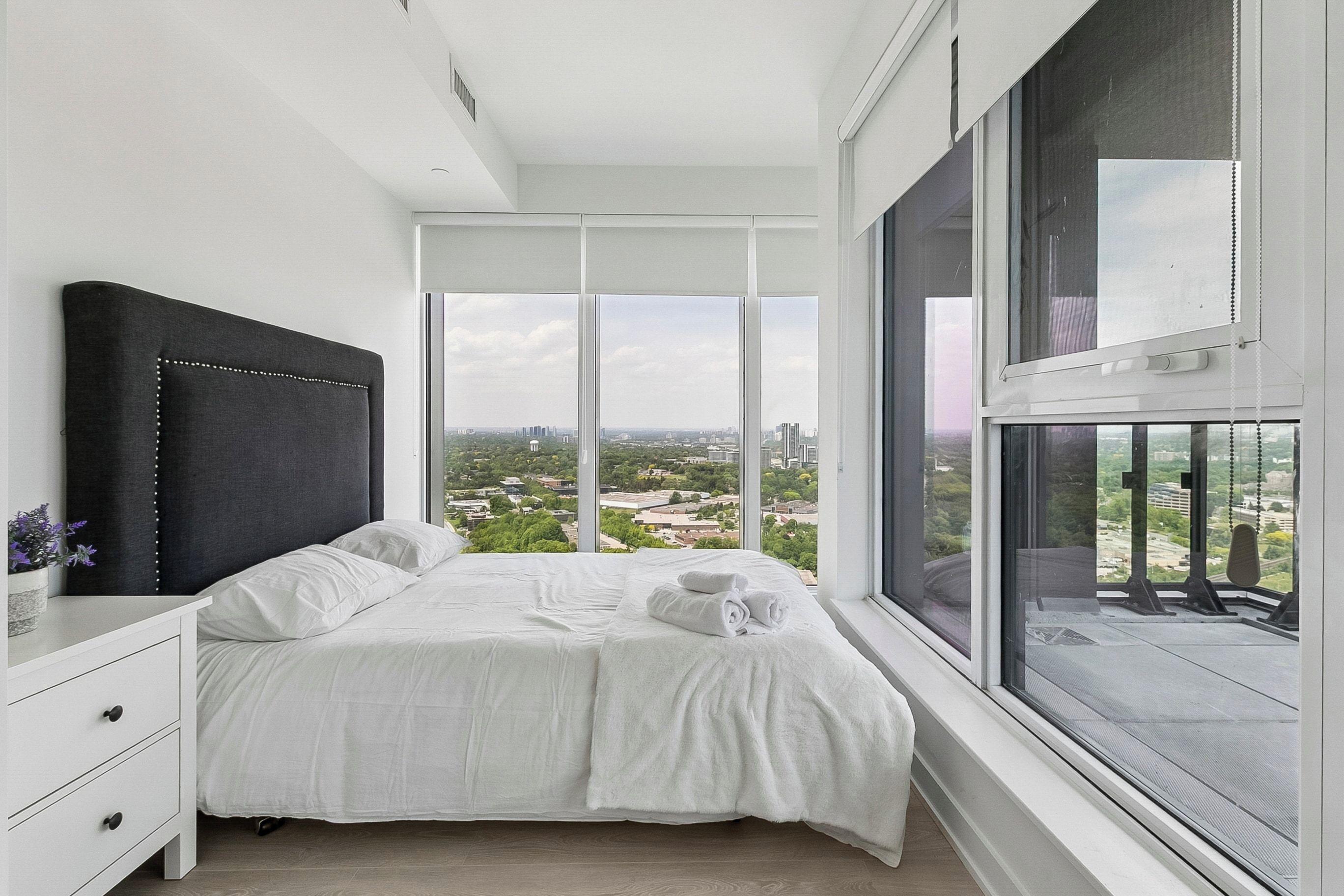A bedroom with a large window, a bed, a nightstand, and a view of the city.