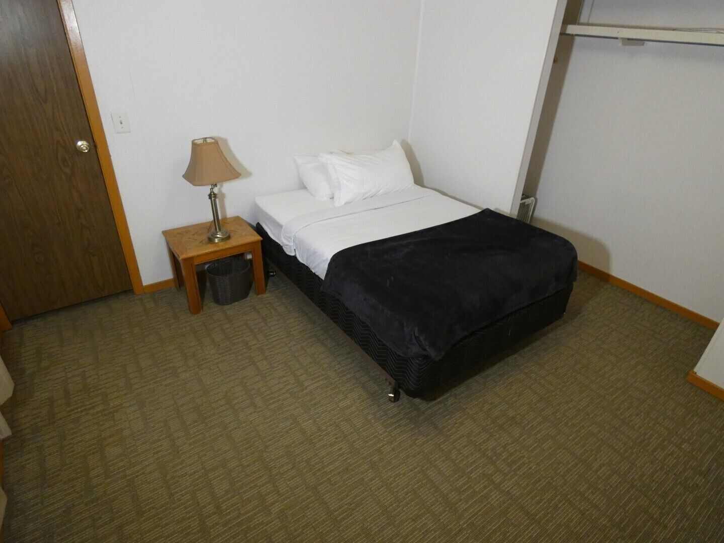 A hotel room with a bed, a nightstand with a lamp, a trash bin, and a wardrobe.