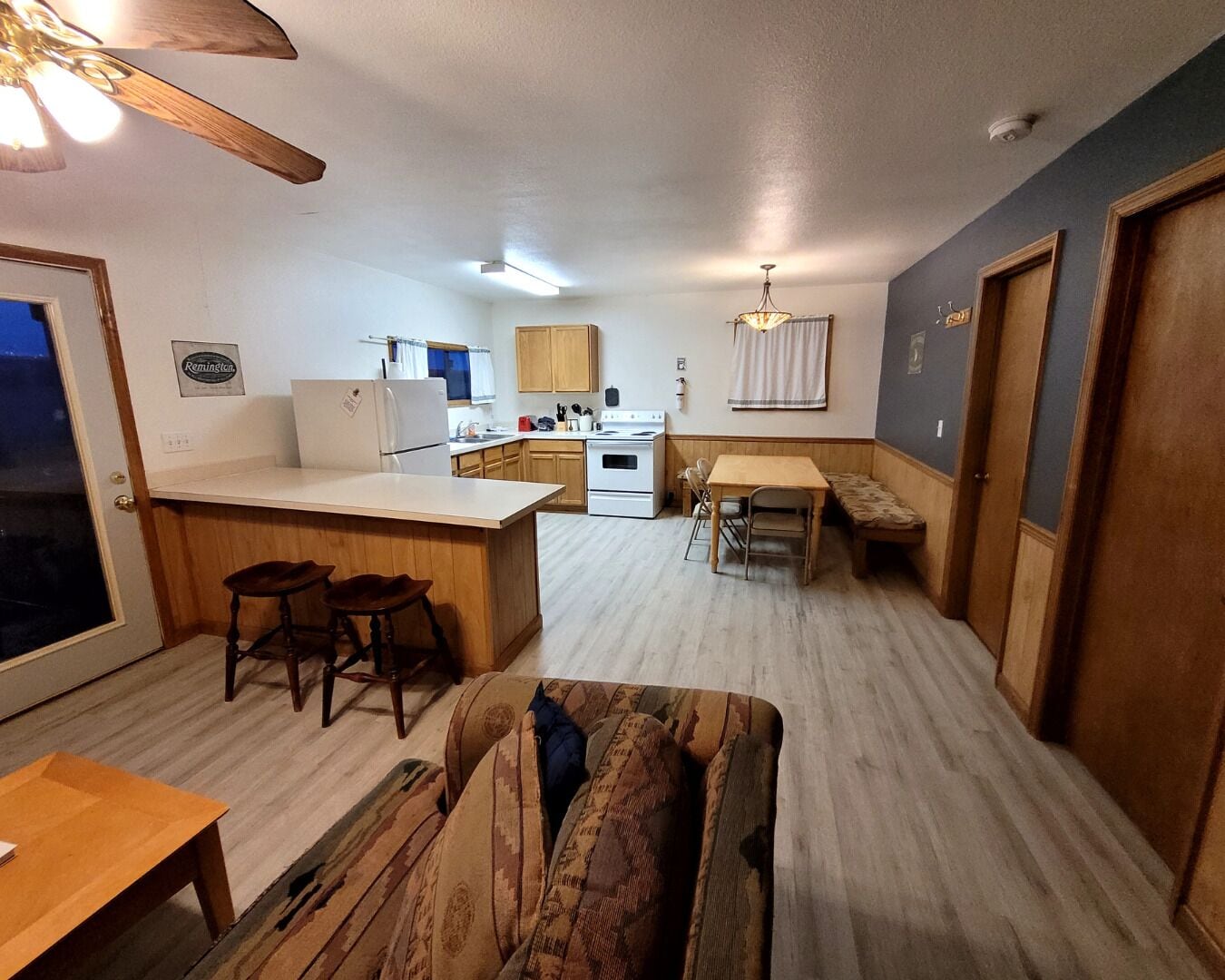 Eine kompakte Küche mit Kühlschrank, Herd und Holzschränken. Es gibt einen Essbereich mit Tisch und Stühlen sowie eine kleine Sitzecke mit einem gemusterten Sofa.