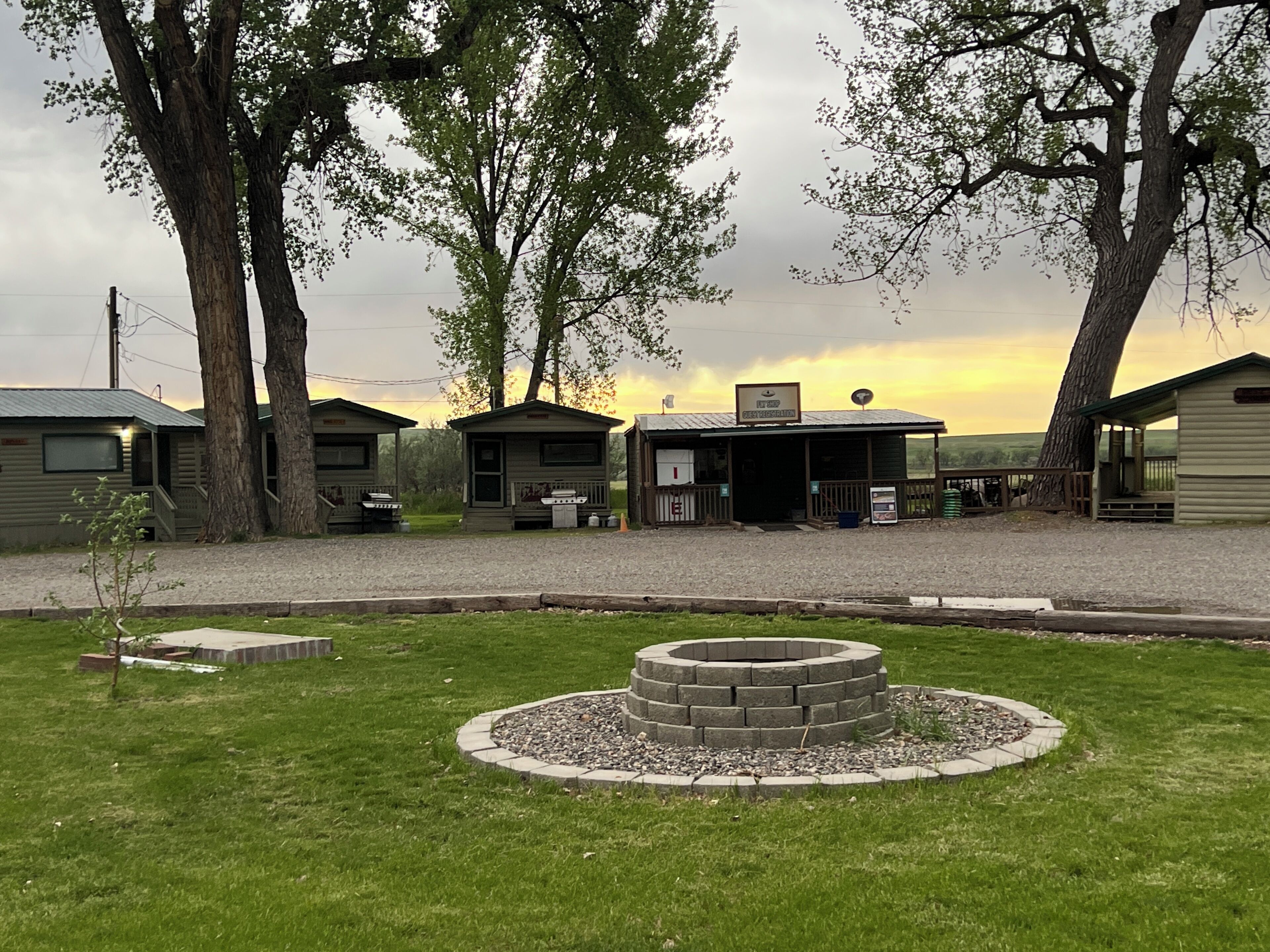 Un terrain de camping comprenant plusieurs cabanes, un foyer central et un sentier de gravier.