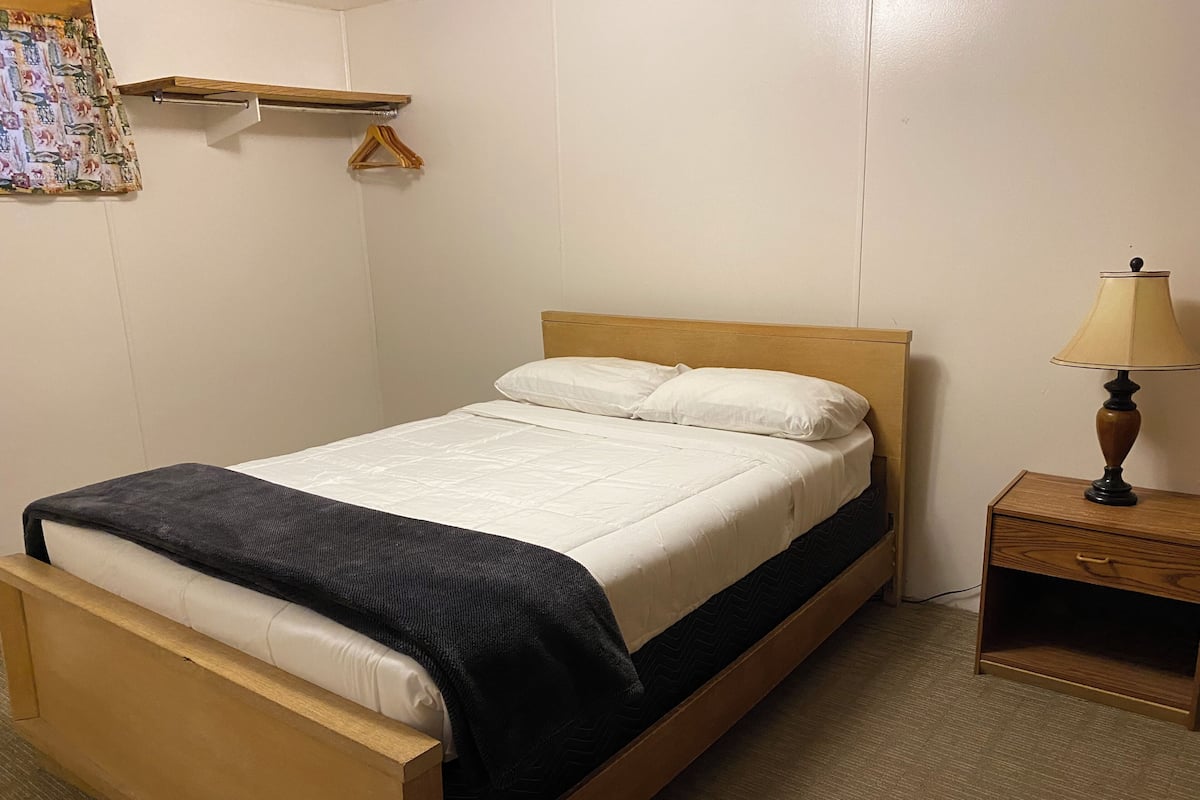 Uma cama de solteiro com estrutura de madeira, uma mesinha de cabeceira com abajur e uma pequena estante com cabides.