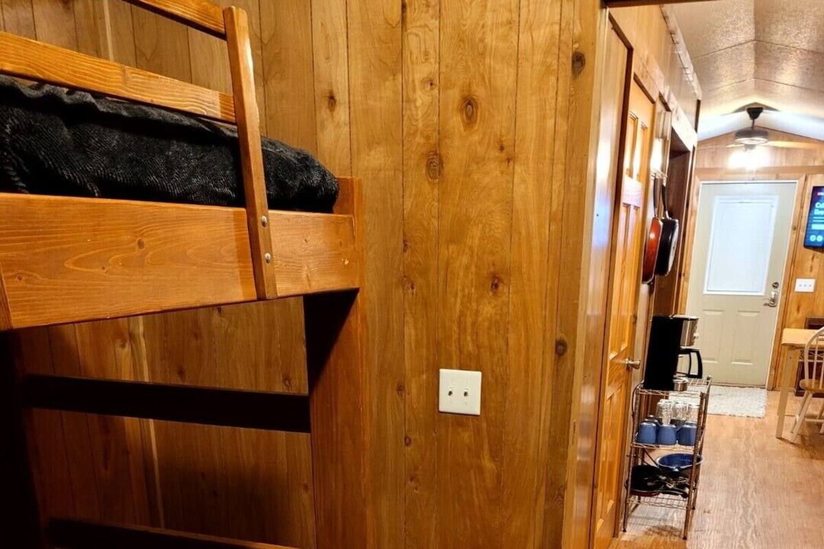 Uma cama beliche com colchão preto, paredes de madeira, uma tomada elétrica branca, uma prateleira de metal com objetos e uma porta ao fundo.