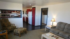 Una sala de estar con sofá, sillón, reposapiés y una mesa de centro. Se ve una cocina al fondo, con armarios y electrodomésticos rojos.