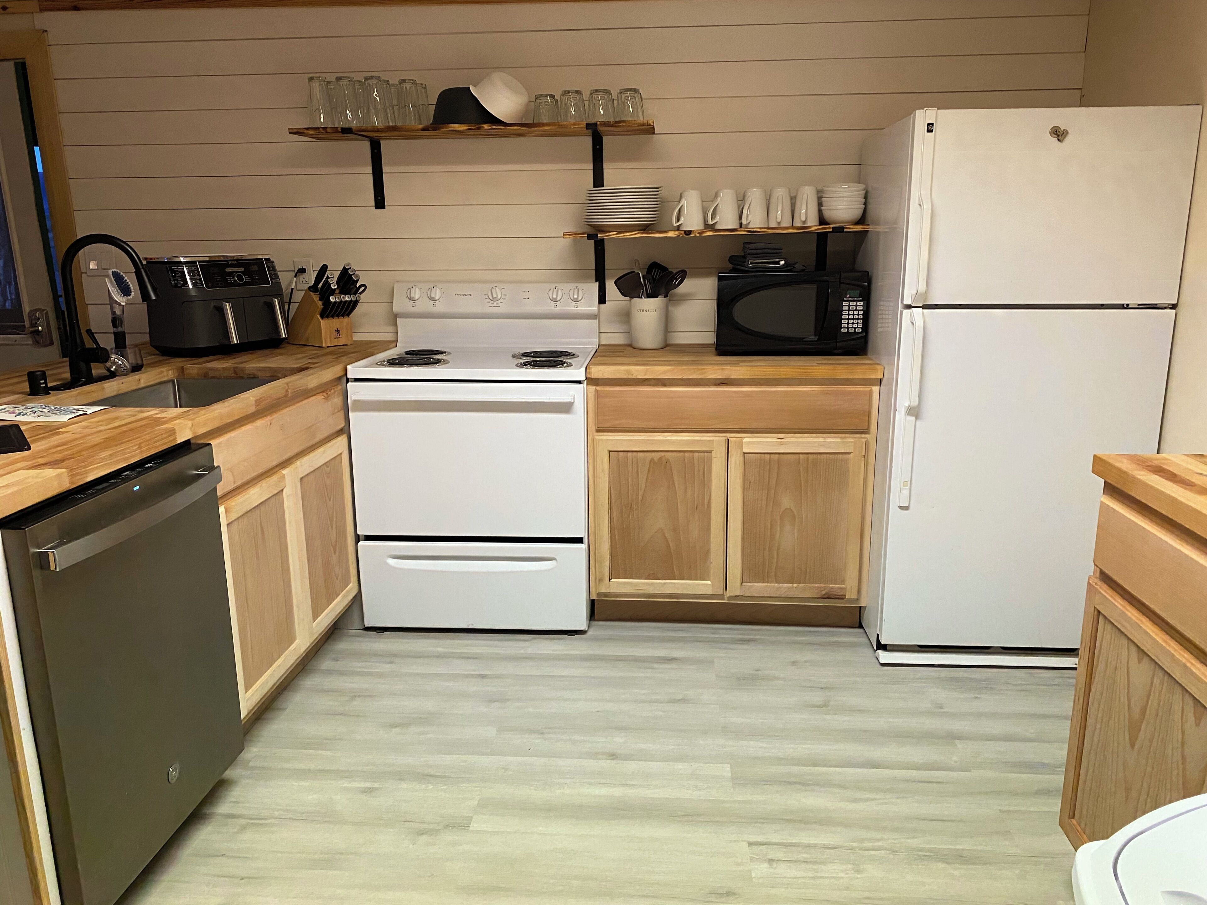 A kitchen with a white stove, microwave, and refrigerator, wooden cabinets, and a shelf with glasses and cups.