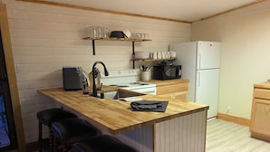 Une cuisine équipée d’un plan de travail en bois, d’une cuisinière, d’un four à micro-ondes et d’un réfrigérateur. On y trouve trois tabourets et une étagère au-dessus du plan de travail.