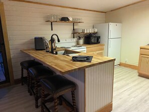A kitchen with a wooden countertop, a stove, a microwave, and a refrigerator. There are three stools and a shelf above the counter.