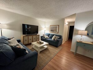 A living room with a TV, two sofas, a coffee table, a lamp, and a framed picture on the wall. - Midtown Beach Condo, Pool, Walk to Beach/Jolly Roger, Linens (Ocean City)