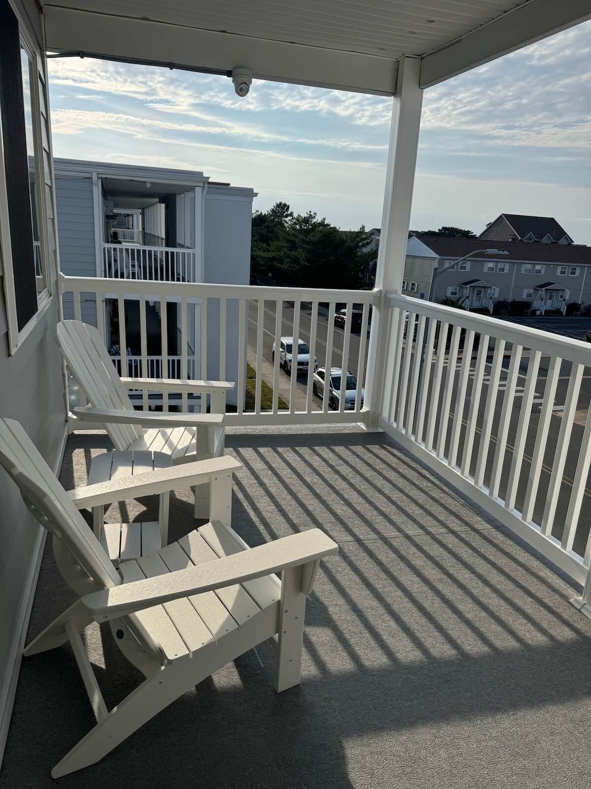 A covered porch with white railings, a white Adirondack chair, and a view of a residential area with houses and parked cars.