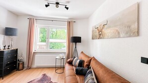 A living room with a brown sofa, a black floor lamp, a ceiling fan, a window with curtains, and a painting of a cow. - Vacation apartment Alte Liebe & Meer (Cuxhaven)