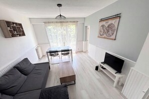En stue med sofa, sofabord, fjernsyn på et stativ, spisebord med stole og et vindue med gardiner.