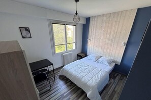 A bedroom with a bed, a window, a radiator, a desk, and a hanging light fixture. - Bright apartment in the city center with balcony (La Roche-sur-Yon)
