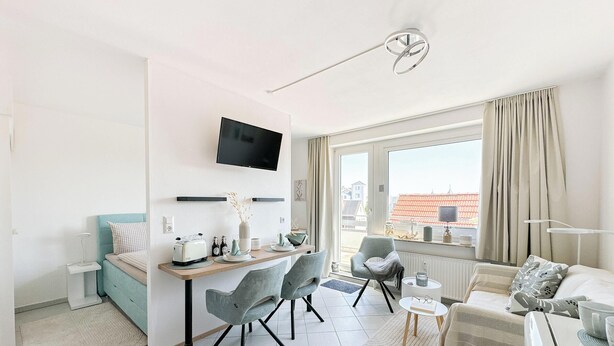 Quarto moderno e bem iluminado, com televisão de tela plana, área de refeições com cadeiras, sofá e uma grande janela com cortinas.