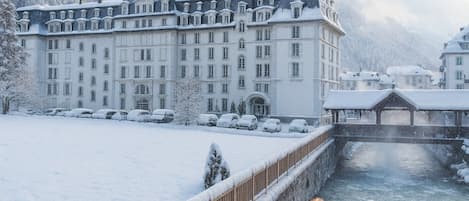 A large, historic building with multiple windows and a snow-covered roof, adjacent to a river.