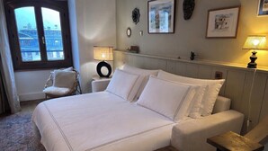Ein Hotelzimmer mit einem großen Bett, einem Sessel, einem Nachttisch, einer Lampe und Blick auf die Stadt durch ein Fenster.