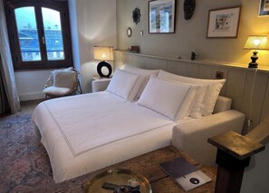 Ein Hotelzimmer mit einem großen Bett, einem Sessel, einem Nachttisch, einer Lampe und Blick auf die Stadt durch ein Fenster.