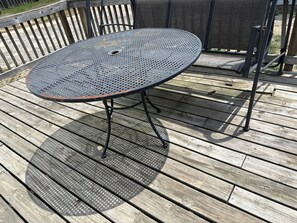 Una mesa redonda de metal con una superficie perforada, sostenida por patas de metal, sobre una terraza de madera.