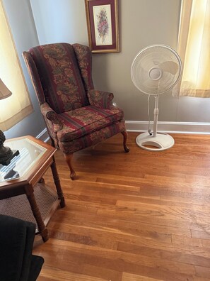 Une pièce comprenant un fauteuil à motifs, un ventilateur sur pied, une table en bois et un tableau encadré accroché au mur.