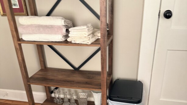 Een houten schapunit met opgevouwen handdoeken en decoratieve objecten, een zwarte prullenbak met doorzichtige zak, en een witte deur op de achtergrond.