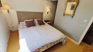 Een netjes opgemaakt bed met een bloemenprint deken en twee kussens, een nachtkastje met een lamp, een spiegel aan de muur, en een deur naar een andere kamer.